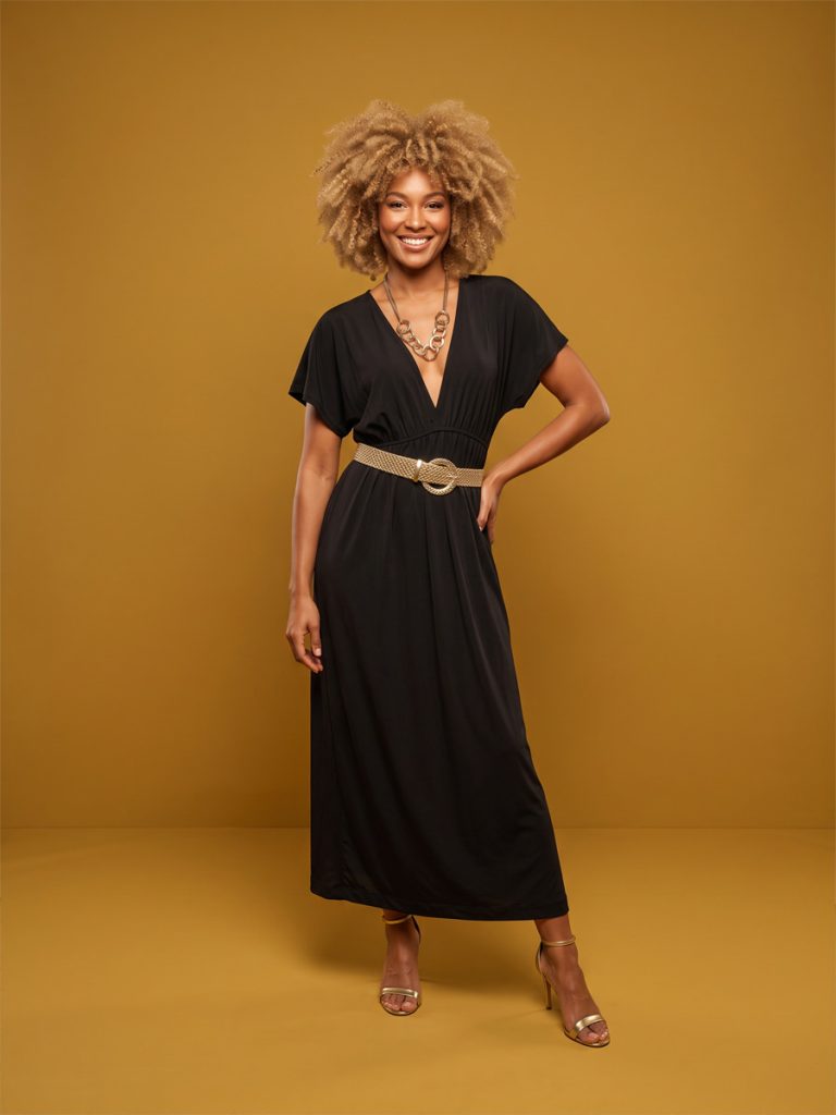 Model is waering a long black 1970s style dress with a gold plait belt an sfro wig